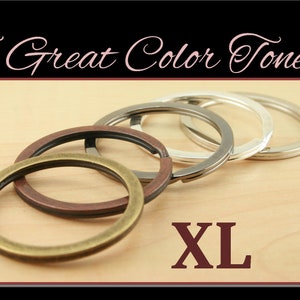 May include: Five large key rings in different metal finishes: antique brass, copper, silver, nickel, and polished steel. The key rings are arranged in a stack on a light wood surface. The text "XL" is printed in red on the wood surface.