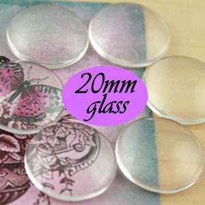 May include: A close-up of six clear glass cabochons, each measuring 20mm in diameter. The cabochons are arranged in a semi-circle on a pink and white patterned surface. The text "20mm glass" is printed in black on a purple background.