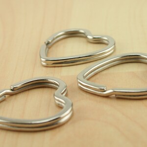 Op de afbeelding: Drie zilveren hartvormige sleutelhangers op een licht houten oppervlak.