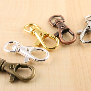 May include: An assortment of metal swivel clasps in various finishes: gold, bronze, silver, and antique brass. These clasps are designed with a spring-loaded gate for secure attachment and are suitable for crafting or jewelry making.