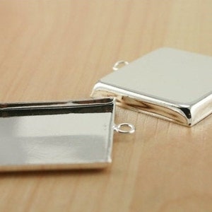 May include: Two silver-toned square pendant trays with open jump rings on each side. The trays are designed to hold a square piece of glass or other material.