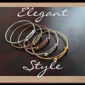 May include: Four silver bangle bracelets with a ball closure. The bracelets are stacked on top of each other. The bracelets are on a brown wooden surface. Elegant Style.