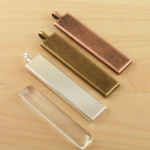May include: Four rectangular metal pendant blanks in silver, brass, and copper finishes. The blanks are all the same size and shape, and they have a small loop at the top for attaching a chain or cord. A clear glass pendant blank is also shown.