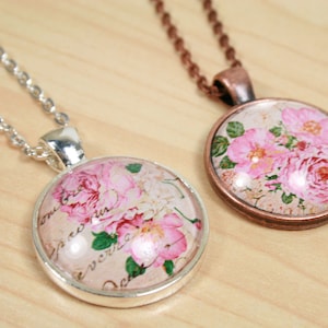 Pode incluir: Dois colares com pingentes redondos de vidro com rosas cor-de-rosa e um design floral vintage. Um colar tem uma corrente de prata e o outro uma corrente de cobre.
