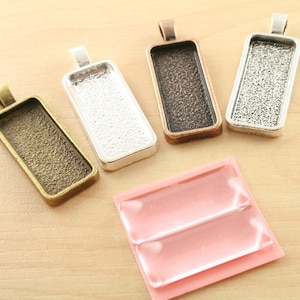 May include: Four silver and bronze pendant trays with a smooth, textured surface. The trays are rectangular and have a loop at the top for hanging. The trays are arranged in a row on a light brown wooden surface.