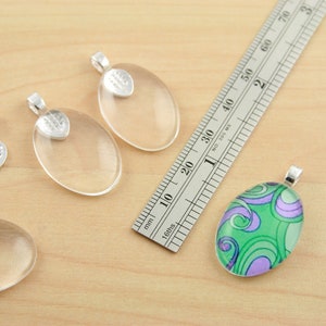 May include: Five clear glass oval pendant charms with silver metal bail caps. One charm has a green and purple swirl design. A ruler is in the background.