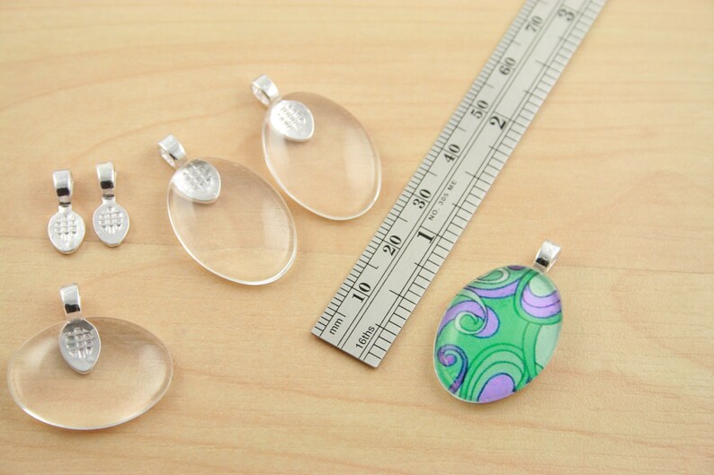 May include: Five clear glass oval pendant charms with silver metal bail caps. One charm has a green and purple swirl design. A ruler is in the background.
