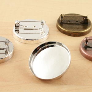 May include: Four round metal bases for making custom brooches. The bases are silver, gold, copper, and blank. Each base has a pin back.