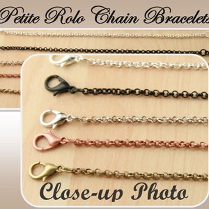 May include: Close-up photo of five petite rolo chain bracelets in silver, black, copper, rose gold, and antique brass. The chains are all the same size and style, but they are different colors. The chains are lying on a wooden surface.
