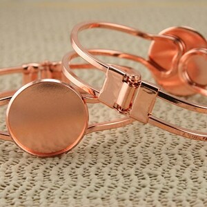 May include: Two rose gold bangle bracelets with a round, flat, rose gold charm attached to each. The charms are blank and appear to be for adding a design.