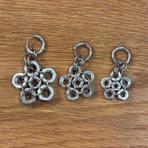 Op de afbeelding: Drie zilverkleurige metalen bedels gerangschikt in afnemende grootte. Elke bedel is opgebouwd uit zeskantige moeren die in een bloemachtig patroon met elkaar zijn verbonden en hangen aan een ronde metalen ring aan de bovenkant van elk stuk.