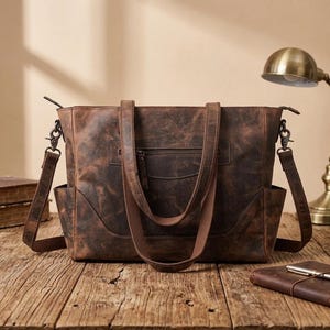 May include: A dark brown distressed leather tote bag featuring two top handles, a detachable shoulder strap with metal clips, a horizontal zippered front pocket, and open side pockets. The bag has a rectangular shape with visible stitching details.