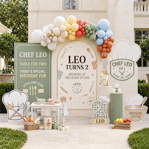 May include: A birthday party display featuring a large arch backdrop with the text LEO TURNS 2, a green sign reading CHEF LEO, and a chef hat sign. Includes a balloon garland, a small table with a basket of bread, and a number 2 sign with a chef hat.