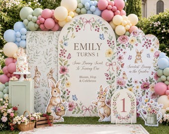 May include: A birthday party backdrop featuring three arched panels decorated with floral illustrations, butterflies, and rabbits. The center panel displays the text EMILY TURNS 1, Some Bunny Sweet Is Turning One, and Bloom, Hop & Celebrate.