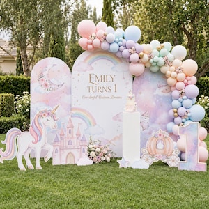 May include: A party backdrop set featuring three arched panels with pastel rainbow, cloud, and moon illustrations. A large balloon garland in pink, blue, and mint surrounds the panels. Cutouts of a unicorn, castle, carriage, and the number 1 are present. Text reads: Emily Turns 1 One-derful Unicorn Dreams.