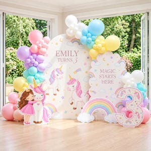 May include: A party backdrop set featuring two arched panels with unicorn, rainbow, and star illustrations, text reading "EMILY TURNS 3" and "MAGIC STARTS HERE," a large number 3 cutout, and a colourful balloon arch in pink, blue, yellow, and purple.