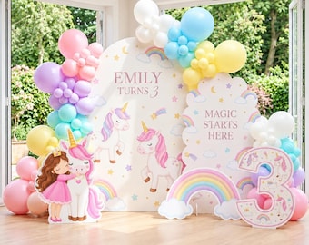 May include: A party backdrop set featuring two arched panels with unicorn, rainbow, and star illustrations, text reading "EMILY TURNS 3" and "MAGIC STARTS HERE," a large number 3 cutout, and a colorful balloon arch in pink, blue, yellow, and purple.
