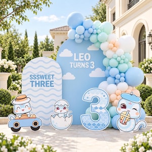 Peut inclure: Un ensemble de décorations de fête avec deux panneaux arqués bleus ornés de nuages blancs et du texte LEO TURNS 3 et SSSWEET THREE. Une grande guirlande de ballons bleus, blancs et pêche entoure les panneaux, avec quatre découpes de serpents.