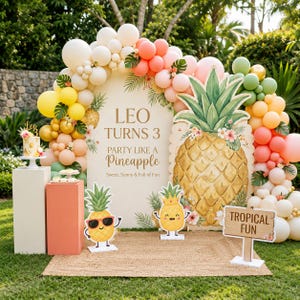 Può includere: Uno sfondo per feste con un grande arco recante il testo LEO TURNS 3 PARTY LIKE A PINEAPPLE, circondato da una ghirlanda di palloncini bianchi, gialli, pesca e verdi, con sagome di ananas e un cartello in legno con la scritta TROPICAL FUN.
