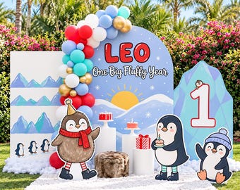 May include: A party backdrop set featuring a blue arch with the text LEO One Big Fluffy Year, a sun graphic, and three penguin cutouts. The display includes a balloon garland, white pedestals with small cakes, and a number one sign.