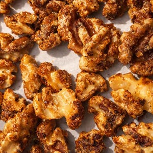 May include: A collection of candied walnut halves coated in a golden-brown, crystalline sugar glaze. The nuts feature a textured, uneven surface with visible caramelized edges and a glossy sheen, scattered across a light-colored, flat surface.