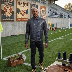 Op de afbeelding: Een man draagt een donkerblauwe jas met slangenpatroon, een zwarte broek en gouden sneakers op een voetbalveld. Achter hem staan posters met de tekst "YES MAN INDUSTRY" en "YMI INDUSTRY". Een houten gereedschapskist en leer staan op de voorgrond.
