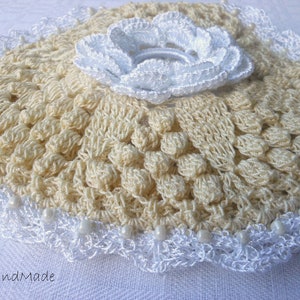 May include: A crocheted doily with a white crocheted flower in the center. The doily is cream colored and has a scalloped edge.