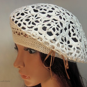 May include: A cream-colored, crocheted beret with an openwork floral design. The beret has a solid band around the base and is adorned with a thin, gold-colored ribbon. The hat is handmade and suitable for various occasions.