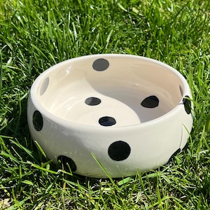 Op de afbeelding: Een ronde, cr&egrave;mekleurige keramische kom met een patroon van zwarte stippen verdeeld over zowel de binnen- als buitenzijde. De kom heeft een gladde, glanzende afwerking en een brede opening met een dikke, afgeronde rand aan de bovenzijde.