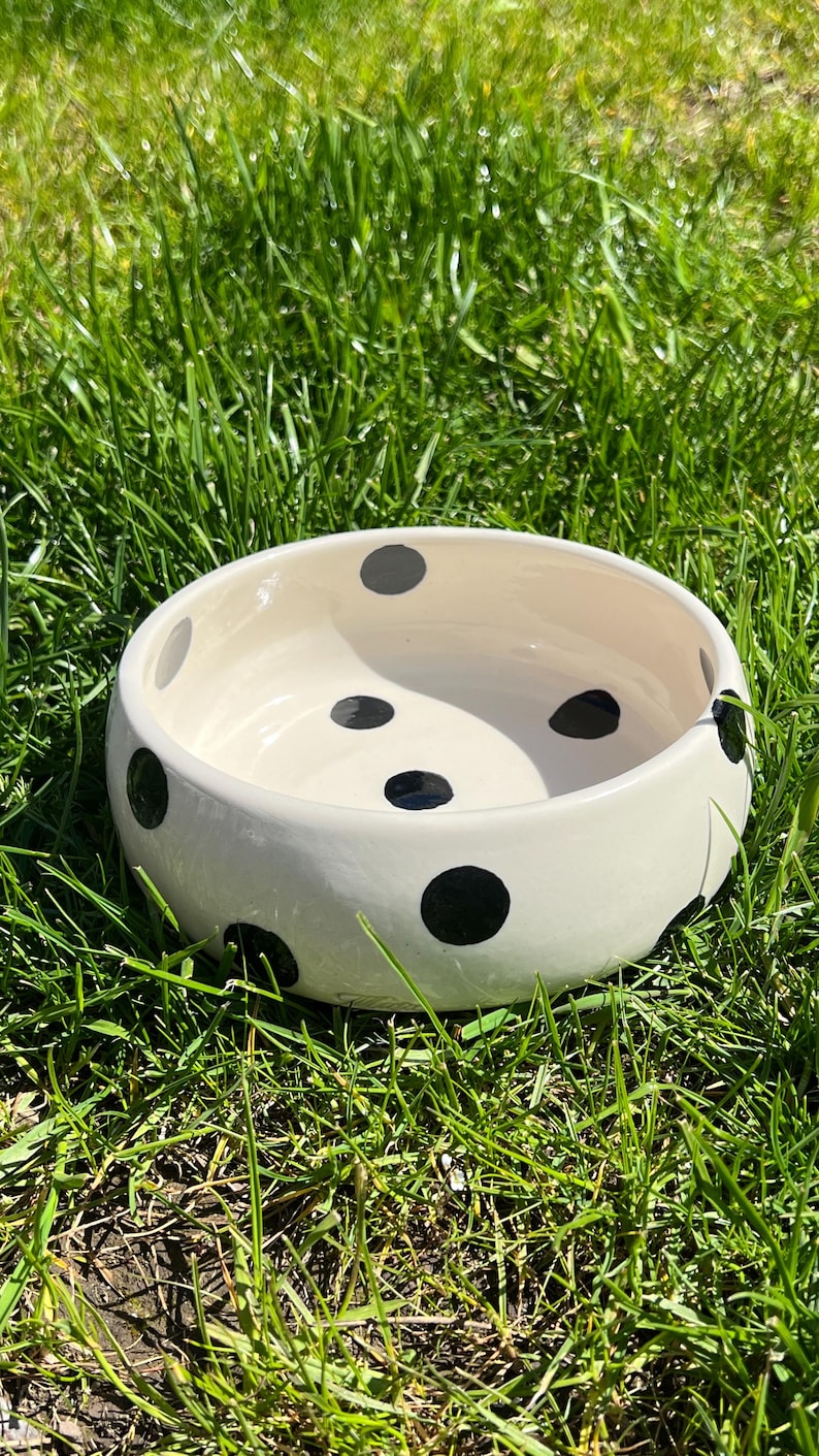 Op de afbeelding: Een ronde, cr&egrave;mekleurige keramische kom met een patroon van zwarte stippen verdeeld over zowel de binnen- als buitenzijde. De kom heeft een gladde, glanzende afwerking en een brede opening met een dikke, afgeronde rand aan de bovenzijde.
