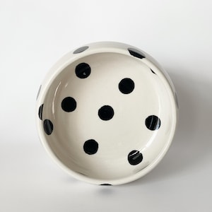 Op de afbeelding: Een ronde keramische kom met een glanzende gebroken witte afwerking, voorzien van een patroon van verspreide zwarte stippen aan de binnenkant en langs de buitenrand. De kom heeft een gladde, gebogen rand en een diepe, open komvorm.