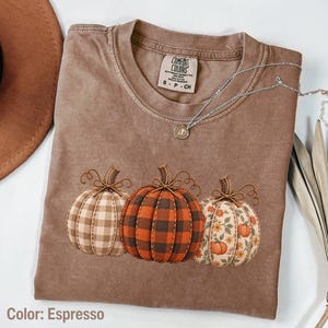 May include: An espresso-colored t-shirt featuring a graphic of three pumpkins. The left pumpkin has a beige gingham pattern, the center has a black and orange buffalo plaid pattern, and the right has a floral print. The shirt has a crew neckline.