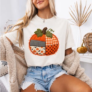 May include: A cream-colored short-sleeve t-shirt featuring a large, central graphic of a patchwork pumpkin. The pumpkin design is composed of various fabric-style patterns including orange gingham, black and white buffalo check, black polka dots, and a white background with small orange pumpkins and black bats.