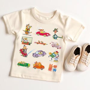 Op de afbeelding: Een crèmekleurig T-shirt met korte mouwen en een kleurrijke grafische print van diverse antropomorfe dieren in auto's, waaronder een kat in een rode auto, muizen in een potloodvoertuig en een vos bij een aambeeld, naast crèmekleurige canvas sneakers.