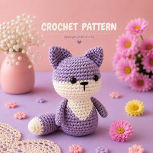 May include: A crochet pattern guide featuring a small, handmade stuffed animal fox. The fox is crafted from light purple and white yarn, with black safety eyes and a stitched nose. The text CROCHET PATTERN STEP-BY-STEP GUIDE is visible.