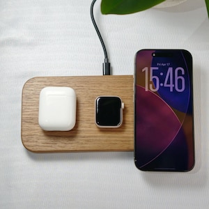 Peut inclure: Station de recharge sans fil en bois avec un étui blanc pour Airpods, une montre connectée noire et un smartphone affichant 15h46. Le chargeur a un câble noir et est posé sur un fond blanc.