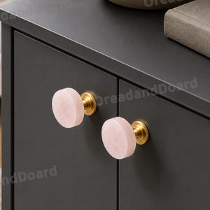 Può includere: Primo piano di un mobile grigio scuro con due pomelli rosa e circolari. I pomelli hanno una base color oro. Una ciotola decorativa e un libro sono visibili sopra il mobile.