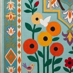 May include: A light blue rug featuring a colorful embroidered pattern of stylized flowers, green stems, and small birds. The border displays a geometric diamond motif with alternating orange and green shapes, alongside small deer figures in the corners.