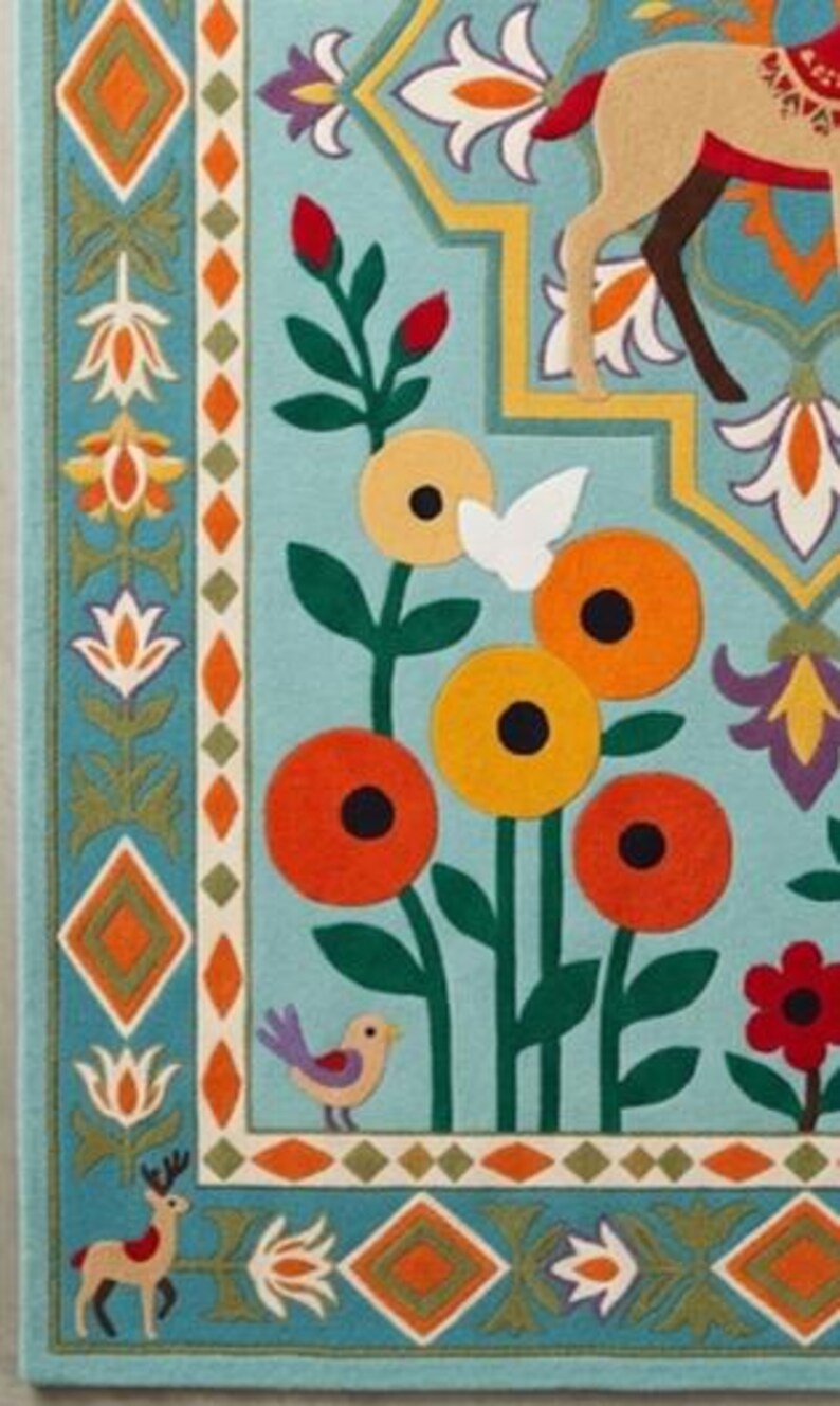 May include: A light blue rug featuring a colorful embroidered pattern of stylized flowers, green stems, and small birds. The border displays a geometric diamond motif with alternating orange and green shapes, alongside small deer figures in the corners.