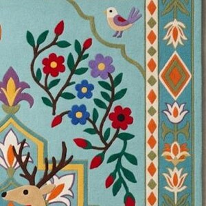May include: A light blue rug featuring a tan deer with dark brown antlers, surrounded by colorful floral motifs, green vines, and a small bird. The border displays a repeating pattern of orange diamonds and white flowers on a blue background.