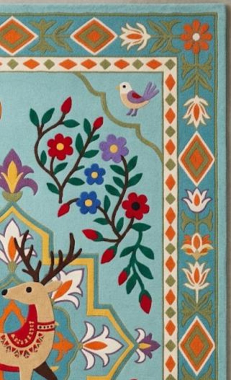 May include: A light blue rug featuring a tan deer with dark brown antlers, surrounded by colorful floral motifs, green vines, and a small bird. The border displays a repeating pattern of orange diamonds and white flowers on a blue background.