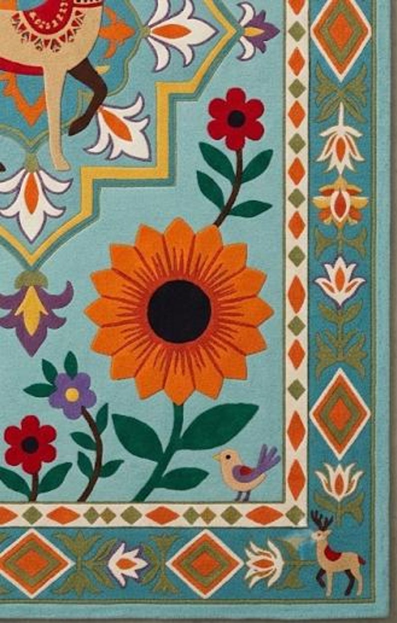 May include: A light blue area rug featuring a large central orange sunflower, smaller red and purple flowers, and green leaves. The design includes a stylized deer, a small bird, and a geometric border with orange diamonds and floral motifs.