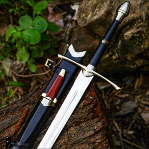 May include: A silver-toned metal sword with a black handle and a matching black scabbard. The scabbard features a small, secondary red-handled knife attached to its side. Both the sword and scabbard have metallic accents and a pointed tip design.