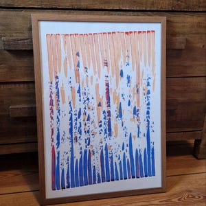 Op de afbeelding: Abstracte kunstafdruk in een houten lijst. Het kunstwerk toont verticale penseelstreken in tinten oranje en blauw tegen een witte achtergrond. De ingelijste kunst wordt tentoongesteld op een houten vloer, leunend tegen een houten dressoir.