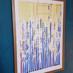 Op de afbeelding: Abstracte kunstafdruk in een houten lijst. Het kunstwerk toont verticale gele en blauwe penseelstreken met rode accenten. Het stuk is gemonteerd op een witte achtergrond en ingelijst in een lichtbruine houten lijst, wat een moderne esthetiek creëert.