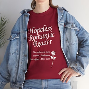 Peut inclure: T-shirt bordeaux avec l'inscription blanche "Hopeless Romantic Reader". En dessous, on trouve "We prefer our men" suivi de "written • Soulmates • Late nights • Slow burn" et une illustration de rose blanche.