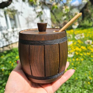Puede incluir: Un pequeño recipiente de madera de color marrón oscuro con forma de barril, tapa a juego y un pomo central. El exterior presenta dos bandas horizontales de cuerda negra y listones verticales. Una cuchara de madera clara está insertada.