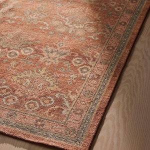 May include: Close-up of a rectangular rug with a distressed appearance and a floral design. The rug has a warm, reddish-orange base colour with faded cream, grey, and yellow floral motifs. A border with a grey and green design frames the edge.