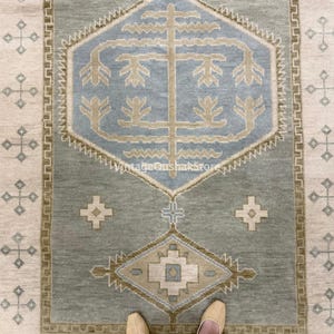 May include: A rectangular area rug with a central geometric design in shades of blue, beige, and green. The rug features a repeating pattern border and a central medallion. The text "VintageOushakStore" is visible on the rug.