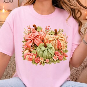 Peut inclure: Un t-shirt rose clair avec un motif central représentant trois citrouilles au crochet orange, pêche et vert. Les citrouilles sont ornées de nœuds, de rubans en dentelle et de petites fleurs au crochet dans des tons rose, jaune et vert.